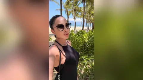Stylish woman in a black bodysuit and sunglasses relaxing on a sunny tropical beach with palm trees.