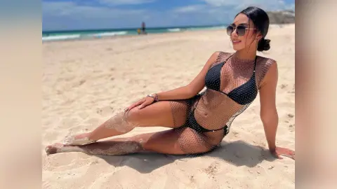 CristalMatthews poses on a sandy beach wearing a black mesh bikini or monokini set.