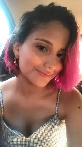 Smiling young woman with pink hair wearing a grey checkered camisole in bright natural light.