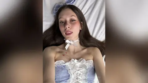 Young woman in blue gingham lace corset and white bow headband lying on white bedding with a soft aesthetic.