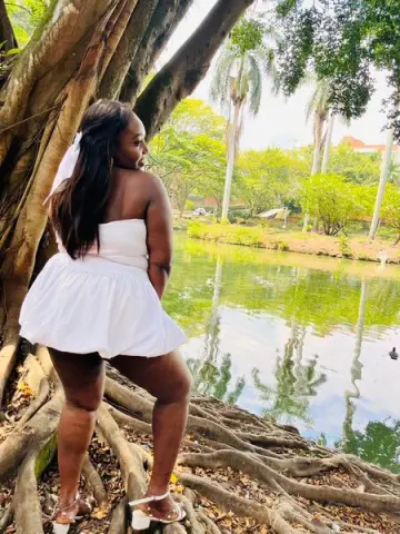 Curvy woman in a white summer romper standing by a tropical lake near large tree roots.