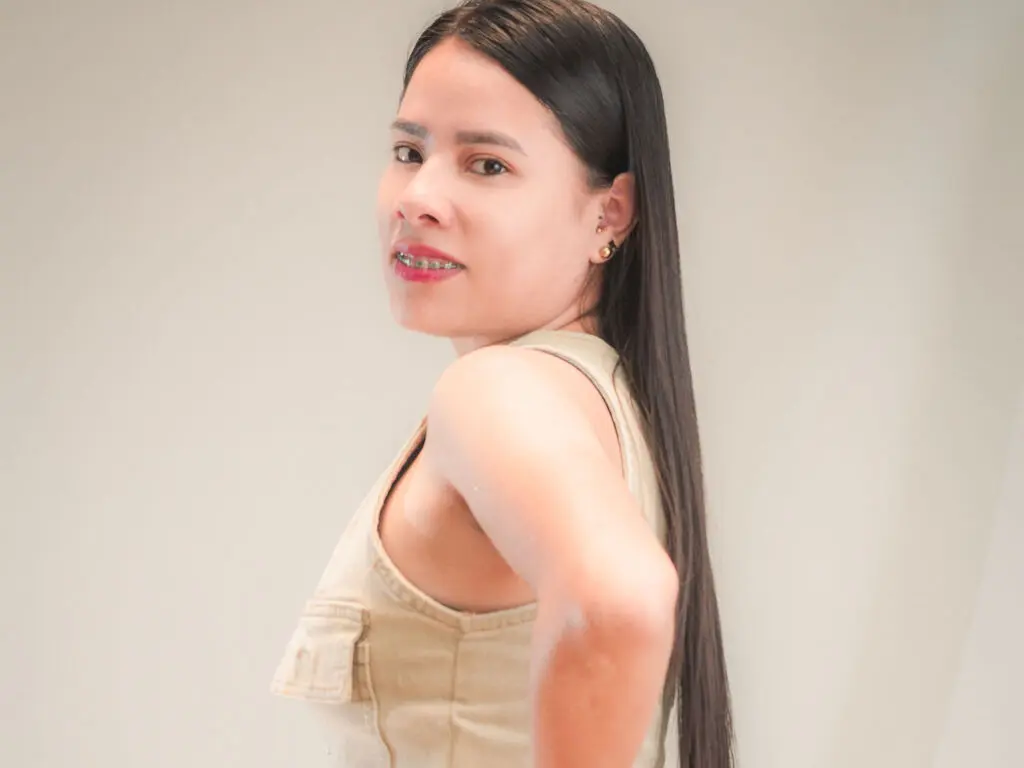 Smiling young woman with long dark hair wearing a beige sleeveless top in a bright minimalist setting.