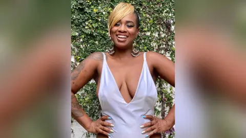 Smiling woman in a white plunging bodysuit posing against a lush green ivy wall.