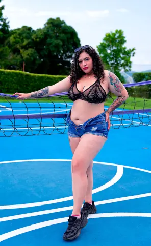 kayllaEvanss poses on a blue court wearing a black lace bra and denim shorts, showing off her tattoos and long curly hair.