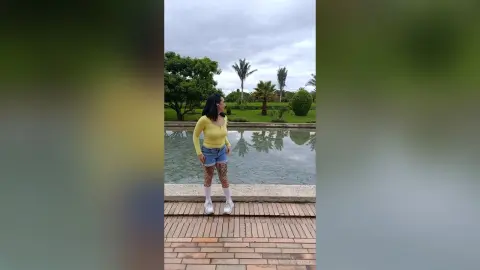 Young woman in a yellow long-sleeve top and denim shorts standing by a serene garden pond under a cloudy sky.