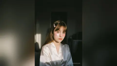 Young woman in white silk pajamas with pink eye patches relaxing in soft, moody indoor lighting.