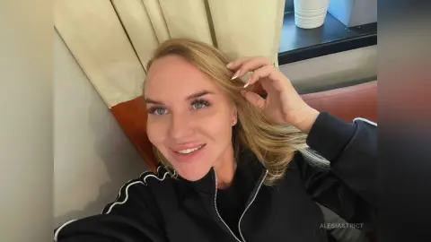 Smiling blonde woman in a black zip-up tracksuit relaxing indoors near a window.