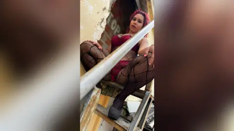 Woman in red bodysuit and fishnet tights sitting on a ladder against a rustic wall in a moody pose.