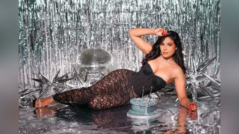 Glamorous woman in a black lace outfit reclining by a disco ball and cake against a silver tinsel backdrop.