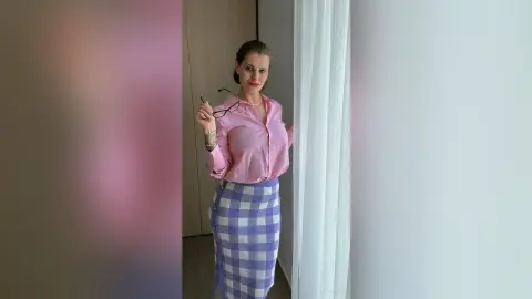 Smiling woman in a pink button-down shirt and lavender gingham skirt standing by a window in a bright room.