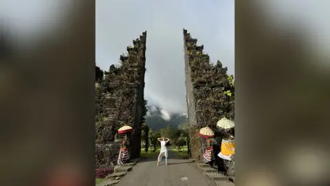 Woman in white summer outfit standing before a massive ancient stone gateway in a misty tropical garden.
