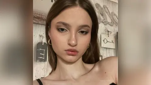 Young woman with natural makeup posing in a rustic room with vintage wall decor.