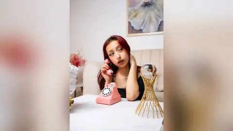 AuroraBellmontk with red hair poses behind a pink rotary phone and a glass crystal ball, wearing a black top in a bright room.