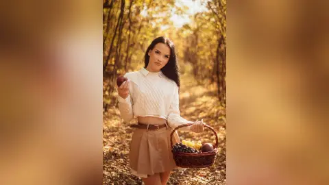 Young woman in a white blouse and tan skirt holding a fruit basket in a sunny autumn forest.