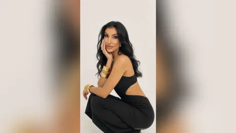 Elegant woman in a black cutout jumpsuit and gold jewelry posing against a white studio background.