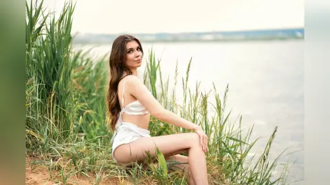 Young woman in white bikini sitting by a lake surrounded by tall green grass in soft natural light.