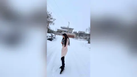 Stylish woman in a beige dress and black boots walking through a snowy residential street on a winter day.