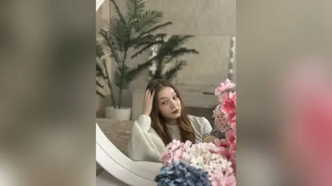 Young woman in a white sweater posing thoughtfully near pink flowers in a bright, minimalist room.