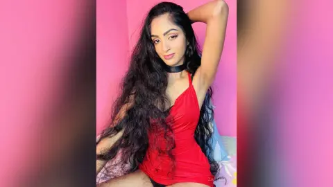 Young woman with long wavy hair in a red tank top and black choker posing against a pink background.