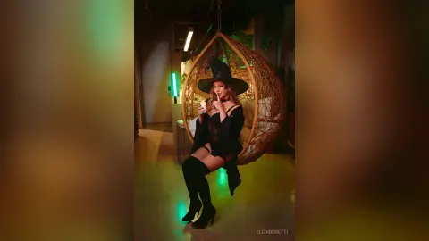 Woman in a witch costume and black boots sitting in a hanging egg chair under moody green lighting.
