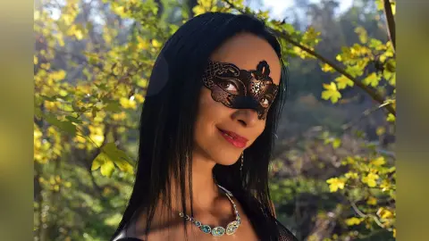 Mysterious woman in a black lace masquerade mask and silver necklace in a sunlit autumn forest.