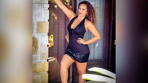 Smiling woman in a black halter bodysuit and sequin mini skirt posing by a stone wall in bright sunlight.