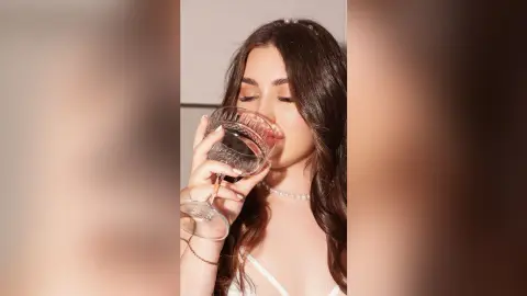 KeiraHarts with long brown hair, wearing a white top and pearl necklace, while drinking from a glass.