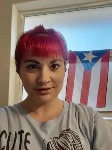 RicanShy looks at the camera with her bright pink hair and a grey t-shirt, with a Puerto Rican flag visible in the background.
