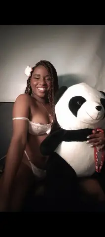 Smiling Black woman in white lace lingerie hugging a large panda plushie in a cozy indoor setting.