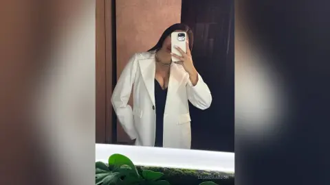 LeonidaAbdon poses in a white blazer over a black top, showing a hint of cleavage while holding a phone for a mirror selfie.