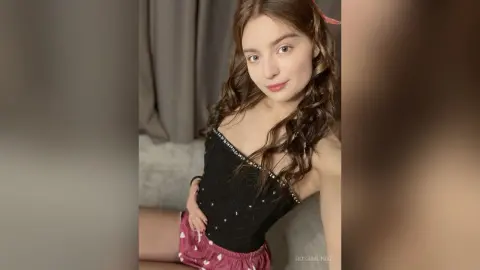Young woman with long wavy hair wearing a black sequined camisole and pink patterned shorts in a soft indoor setting.