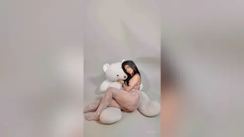Young woman in lace lingerie hugging a giant white teddy bear in a cozy, minimalist studio setting.