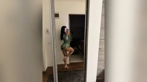 AlmaKane — Young woman in a green floral mini dress posing for a mirror selfie in a modern minimalist room.