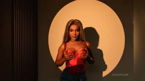 Glamorous woman in a red corset top posing against a dramatic circular shadow in a moody studio setting.