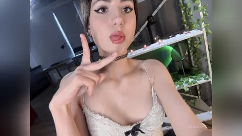 AriaLush poses in a white lace camisole, sticking her tongue out while making a peace sign near her neck.