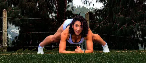 Athletic woman in white fitness wear performing a yoga stretch on green grass in a lush outdoor setting.
