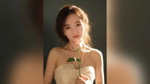 Elegant woman in a white strapless dress holding a single pink rose in soft, warm lighting.