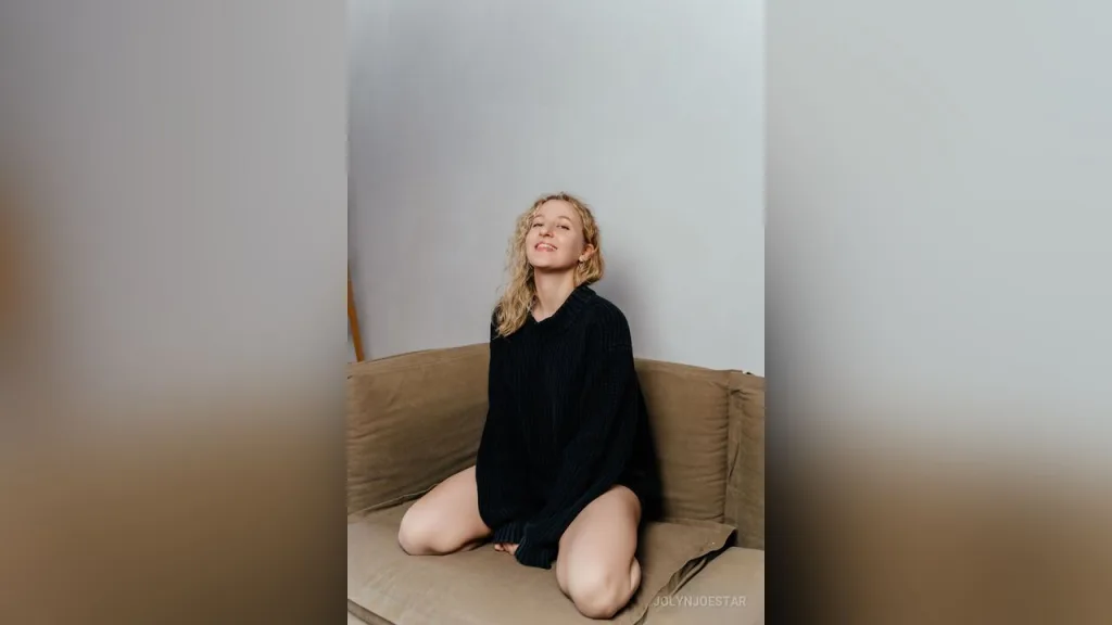 JolynJoestar sits on a brown sofa wearing a black slip dress with a high slit, showing her bare legs.
