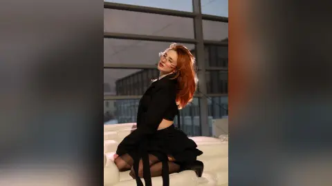 AlbertaCorrell is a redhead in a black outfit sitting on a white couch in front of a large window.