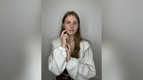 CatherynBuckland poses against a plain grey wall, wearing a white hooded sweatshirt and a small necklace, touching her cheek with one hand.