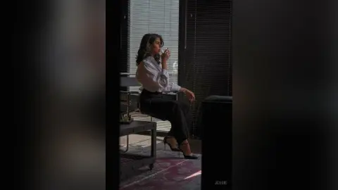 Elegant woman in white blouse and black trousers sipping coffee in a moody, sunlit modern office setting.
