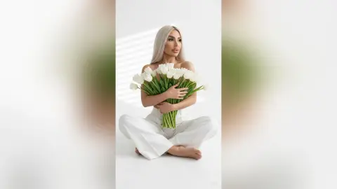 Blonde woman in white trousers holding a large bouquet of white tulips in a bright, minimalist setting.