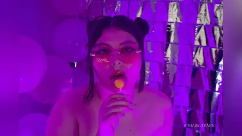 AnnaBeltrann licks a yellow lollipop while wearing pink glasses, posing against a background of white balloons and silver tinsel under purple light.
