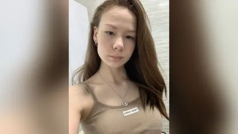 Young woman with long brown hair wearing a beige ribbed tank top in a bright indoor setting.