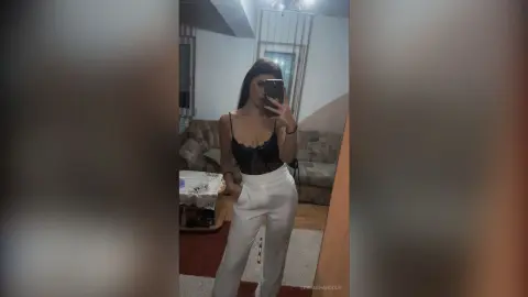 Young woman in a black lace camisole and white high-waisted trousers taking a mirror selfie in a cozy living room.