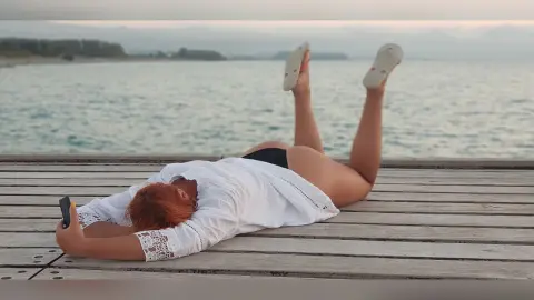AryaSin lies on her stomach on a wooden pier, wearing a white lace shirt and black panties, looking at her phone with her legs raised in the air.