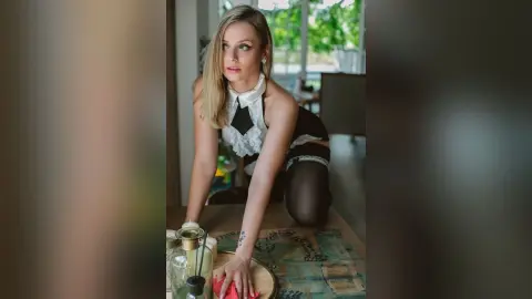Gia poses kneeling on a table in a black and white lace outfit, looking off to the side with her blonde hair down.