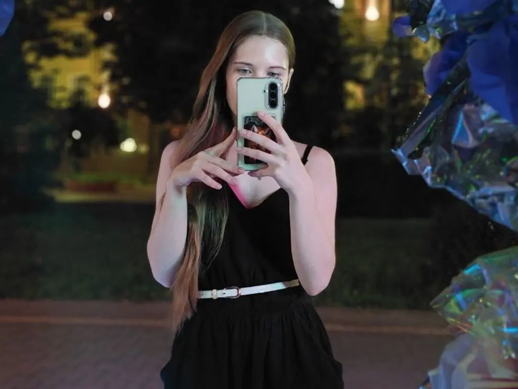Young woman in a black dress taking a mirror selfie outdoors at night with festive lights.