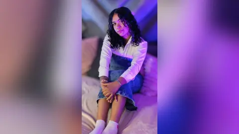 NaomiVelour sits on a bed wearing a white long-sleeve shirt and a denim skirt, looking at the camera with her dark curly hair down.