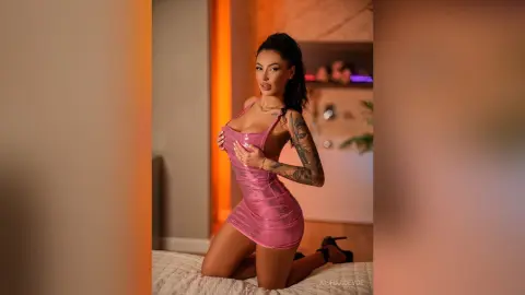 Glamorous woman in a tight pink satin mini dress posing on a bed in warm, moody indoor lighting.